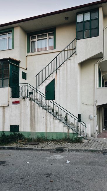 A multi-storey residential building exterior during the home relocation process, showing an external metal staircase with black railings leading from the pavement to a rear door on the first floor. Th