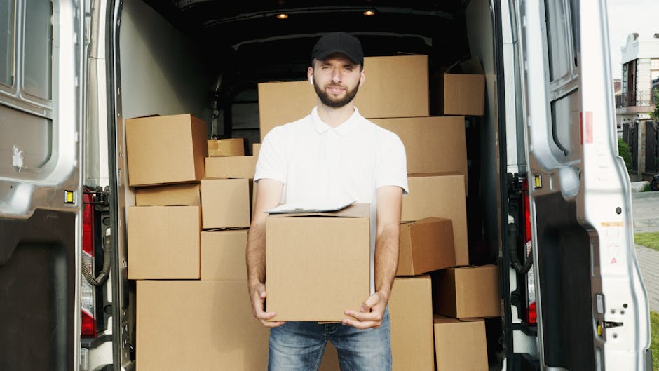 A man dressed in a white polo shirt, jeans, and a black cap is standing at the rear of an open moving van, holding a medium-sized cardboard box during a house relocation. The van’s interior is filled with stacked cardboard boxes of various sizes, some wrapped in plastic or packing materials, ready for transport. The loading process is taking place outdoors on a pavement with nearby residential buildings. A trolley and packing blankets are visible inside the van, supporting the furniture transport and packing activities associated with professional removals by Ealing Movers. The scene captures the logistics of loading and organizing household items in preparation for a home move, showcasing the steps involved in a professional furniture transport and packing service in Ealing W5.