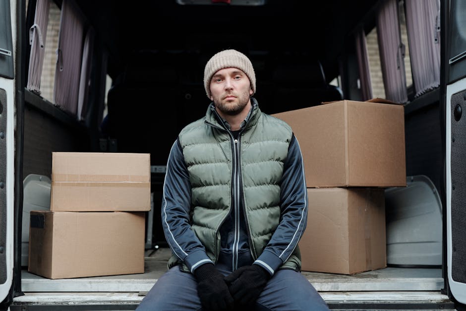 A young man with a serious expression, wearing a grey beanie, a green and navy puffer jacket, and black gloves, sitting inside the open back of a van used for home relocation or furniture transport. Behind him are several cardboard boxes of varying sizes, some sealed with packing tape, placed on the van’s floor and against the side panels. The interior of the van is visible, showing a dark backdrop with a partially open side door and a loading area outside. The scene captures the loading process of packing and moving boxes during a house removal, with the van prepared for transport. Ealing Movers, a professional removal service, is referenced indirectly through the context of house removals and logistics associated with the service category.