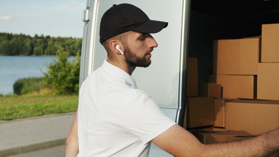 A man dressed in a white polo shirt, jeans, and a black cap is standing at the rear of an open moving van, holding a medium-sized cardboard box during a house relocation. The van’s interior is filled with stacked cardboard boxes of various sizes, some wrapped in plastic or packing materials, ready for transport. The loading process is taking place outdoors on a pavement with nearby residential buildings. A trolley and packing blankets are visible inside the van, supporting the furniture transport and packing activities associated with professional removals by Ealing Movers. The scene captures the logistics of loading and organizing household items in preparation for a home move, showcasing the steps involved in a professional furniture transport and packing service in Ealing W5.