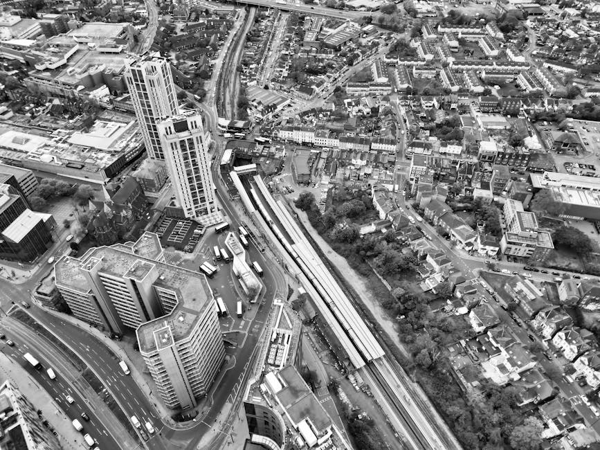 Black and white aerial photograph of an urban area showing a dense cluster of high-rise and mid-rise buildings, with roads and railway tracks running through the scene. The railway tracks are positioned centrally, with multiple trains visible, and are surrounded by commercial and residential structures. A tall modern building is prominent near the bottom left, with other lower-rise office buildings and apartment blocks nearby. Roads are busy with moving vehicles, and the area includes pavement streets lined with trees and smaller residential houses. The image captures an extensive view of the cityscape, highlighting the complexity of urban infrastructure and architecture. This aerial view could relate to city logistics involved in home relocation and furniture transport, similar to services provided by Ealing Movers for house removals and moving logistics in the area around Ealing Broadway station.