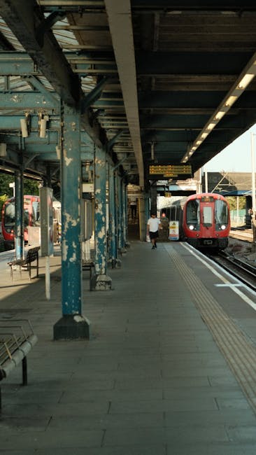The image shows a train station platform during daytime with a red passenger train stopped on the tracks to the right. The platform features a series of blue metal columns supporting an overhead canopy, which has visible signs and lighting fixtures attached. The surface of the platform is paved with concrete and has tactile paving strips along the edge for safety. A single person dressed in casual clothing is walking along the platform toward the train, carrying a small bag. There are additional benches and signs along the platform, and the scene appears to be in a semi-urban setting, suggesting a busy commuter station. This setting is relevant to the context of house removals and moving logistics, illustrating a typical transit environment where furniture transport and home relocation activities might involve coordination with local transit services, as operated by companies like Ealing Movers.